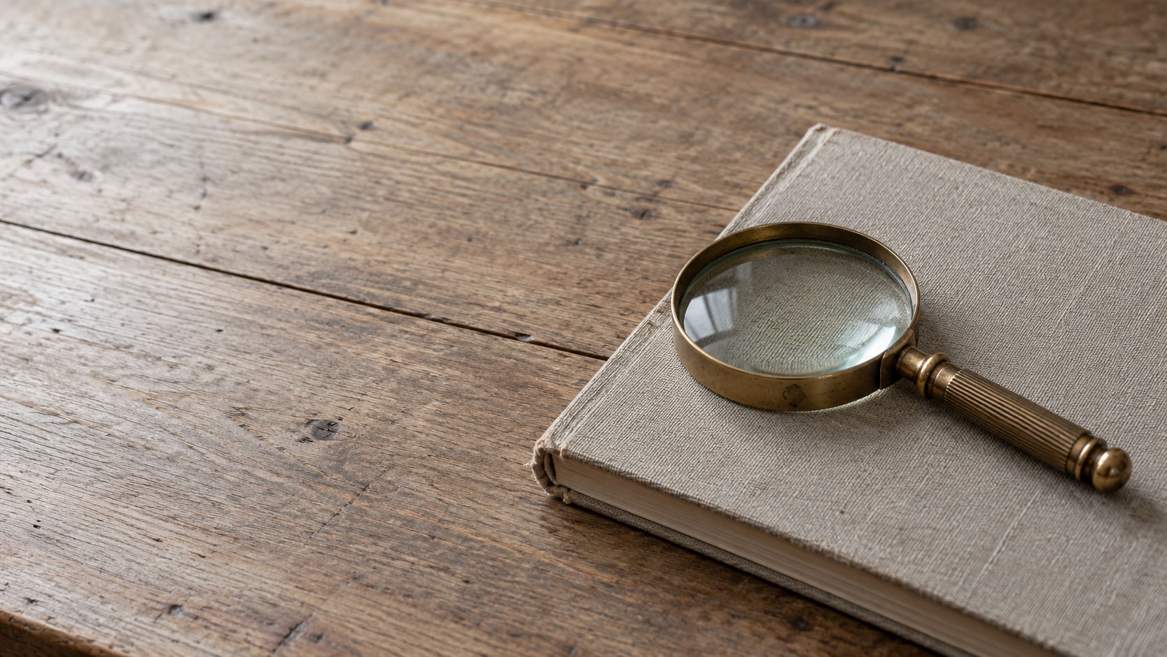 A brass magnifying glass resting on a closed linen-covered notebook on a plain wooden desk.