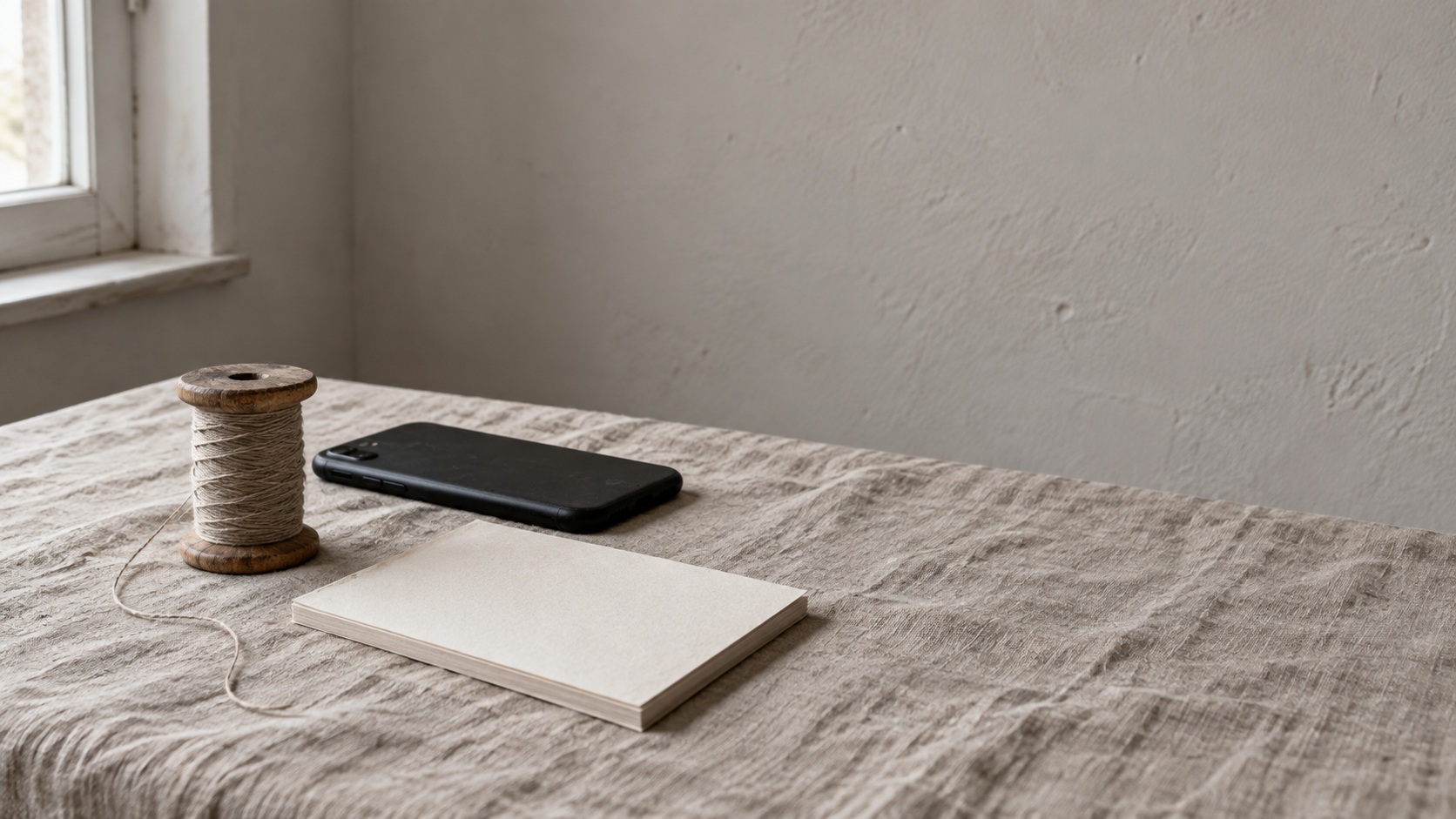Un carrete de hilo junto a un teléfono boca abajo y una libreta en blanco sobre un mantel de lino.