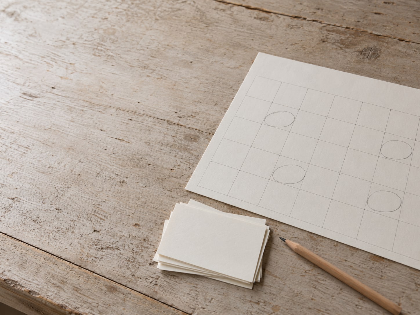 Una hoja de calendario de papel con varios días marcados con lápiz, junto a una pequeña pila de tarjetas en blanco y un lápiz sobre una mesa de madera.