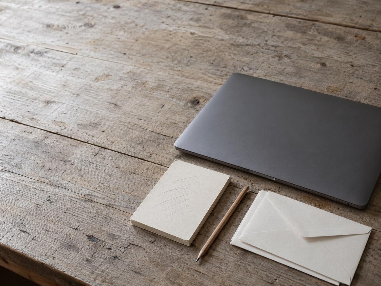 Una laptop cerrada junto a un bloc de notas con marcas de lápiz y una pequeña pila de sobres sin abrir sobre un escritorio de madera gastado.