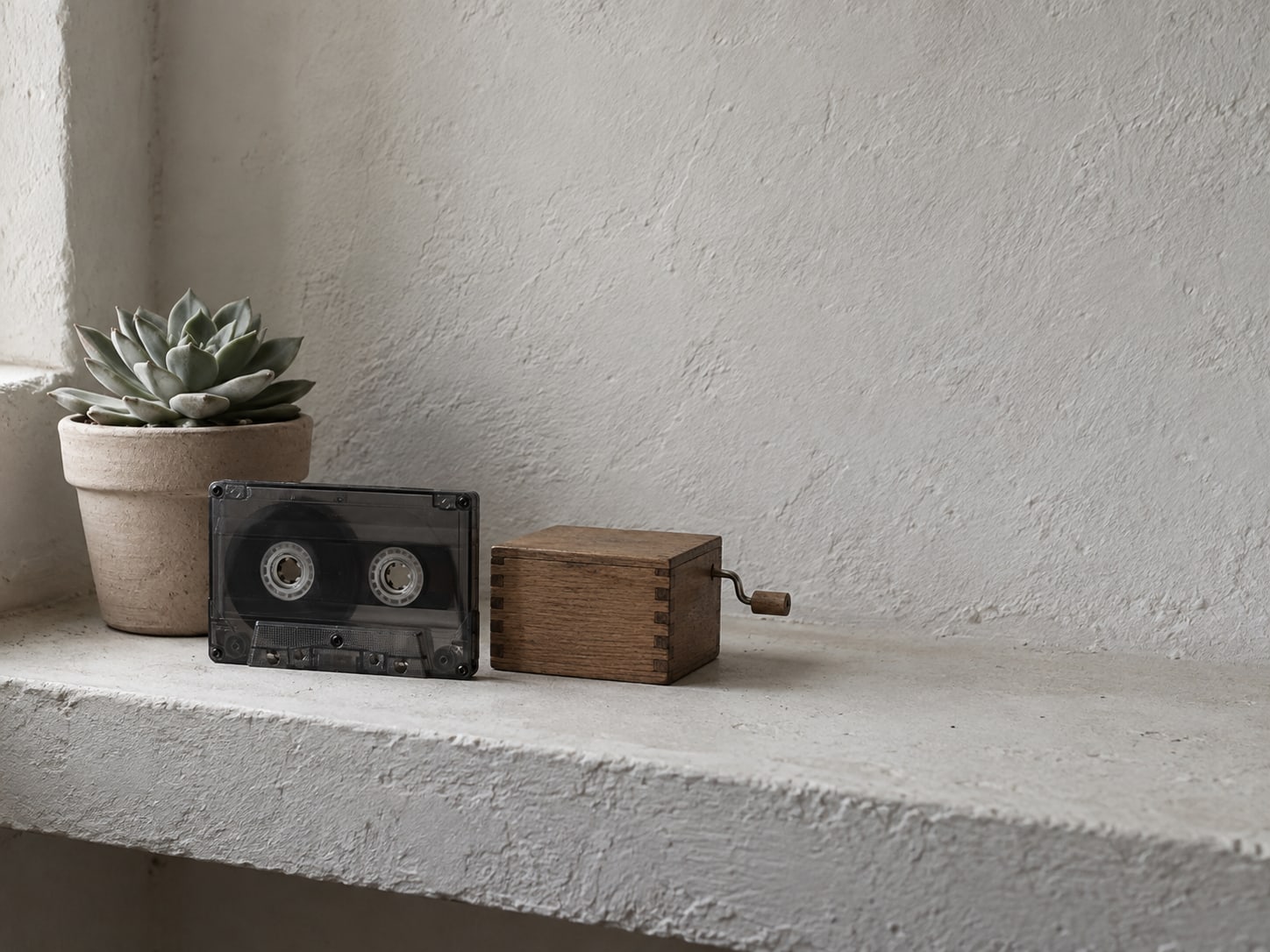 A single reel of cassette tape and a plain wooden music box sit on a clean shelf next to a small potted succulent.
