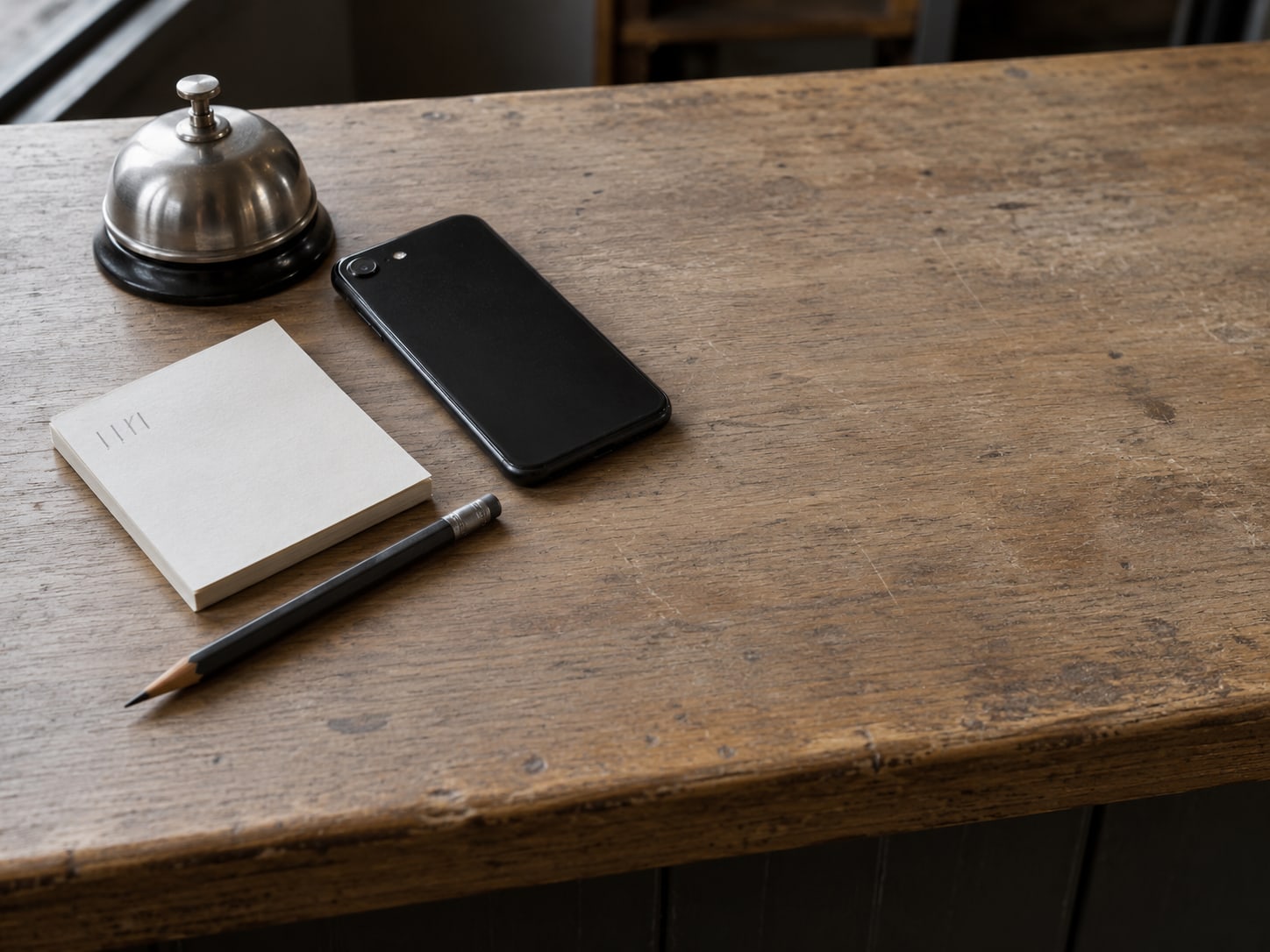 A small paper notepad with pencil tally marks beside a closed phone and a plain counter bell on a shop counter.