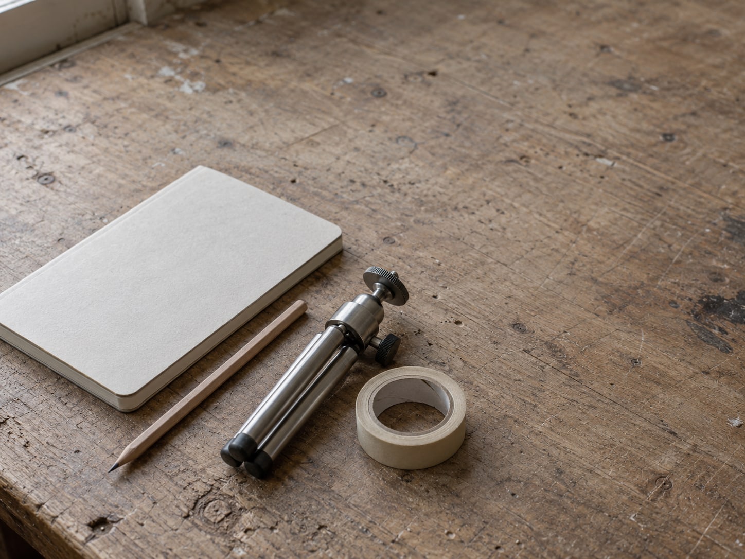 A closed notebook, a pencil and a small tripod stand lie on a workshop bench beside a roll of tape.