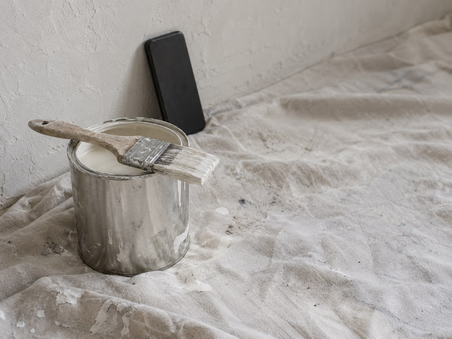 A paintbrush resting across an open paint can on a drop cloth, with a phone propped face down against a wall.
