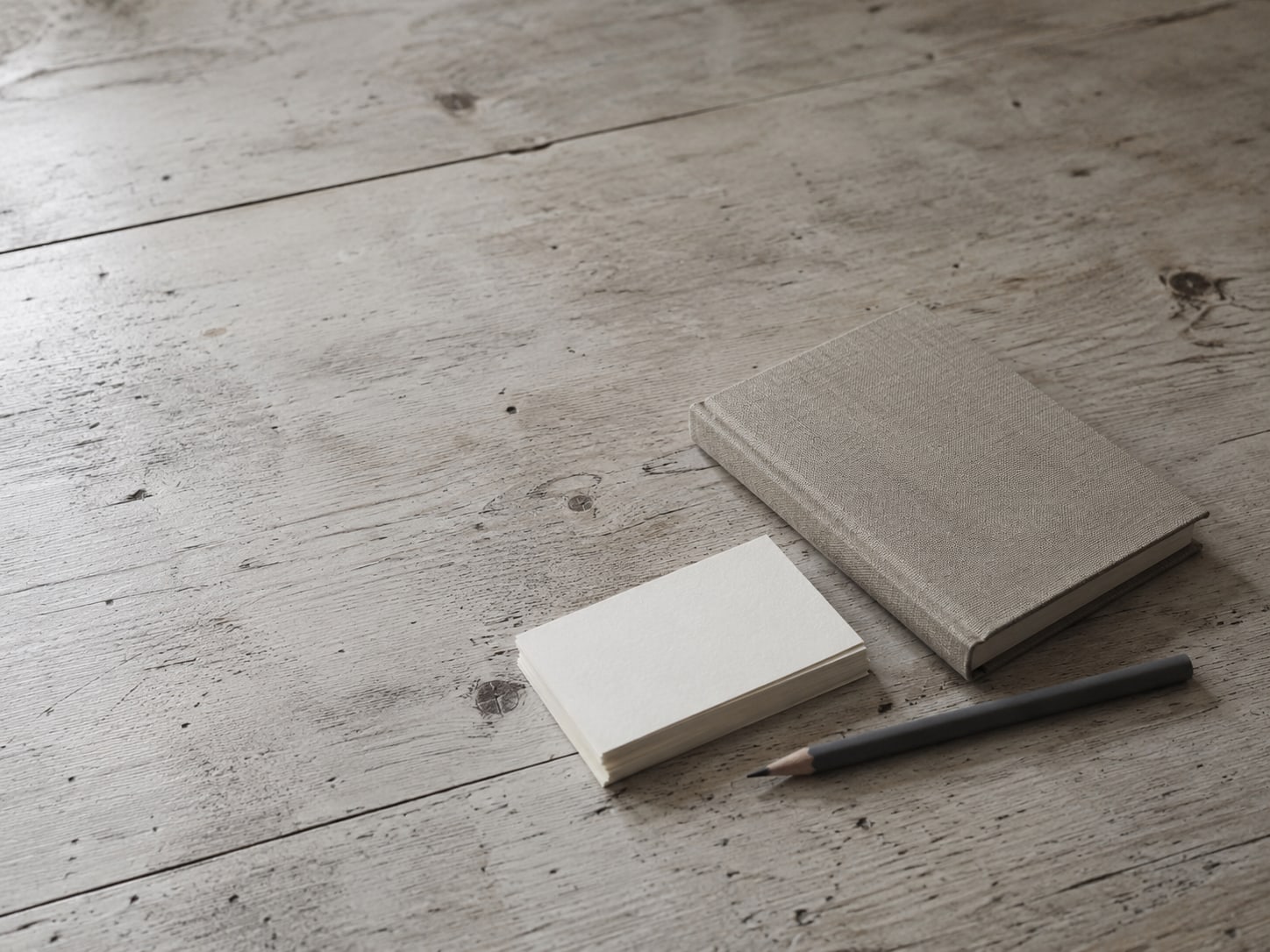 A closed notebook and a pencil lie beside a small stack of blank index cards on a worn wooden desk.