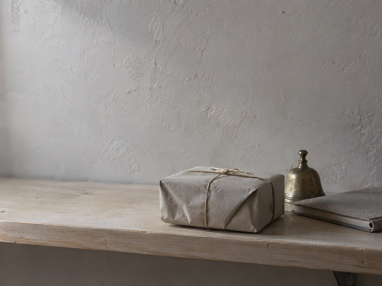 A single wrapped parcel sits on a bare wooden shelf beside a small brass bell and a closed notebook.