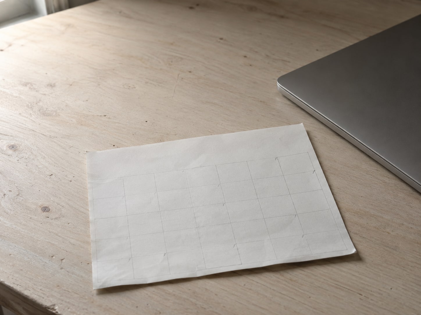 A paper calendar page with small unreadable pencil ticks sits next to a closed laptop on a pale wooden table.