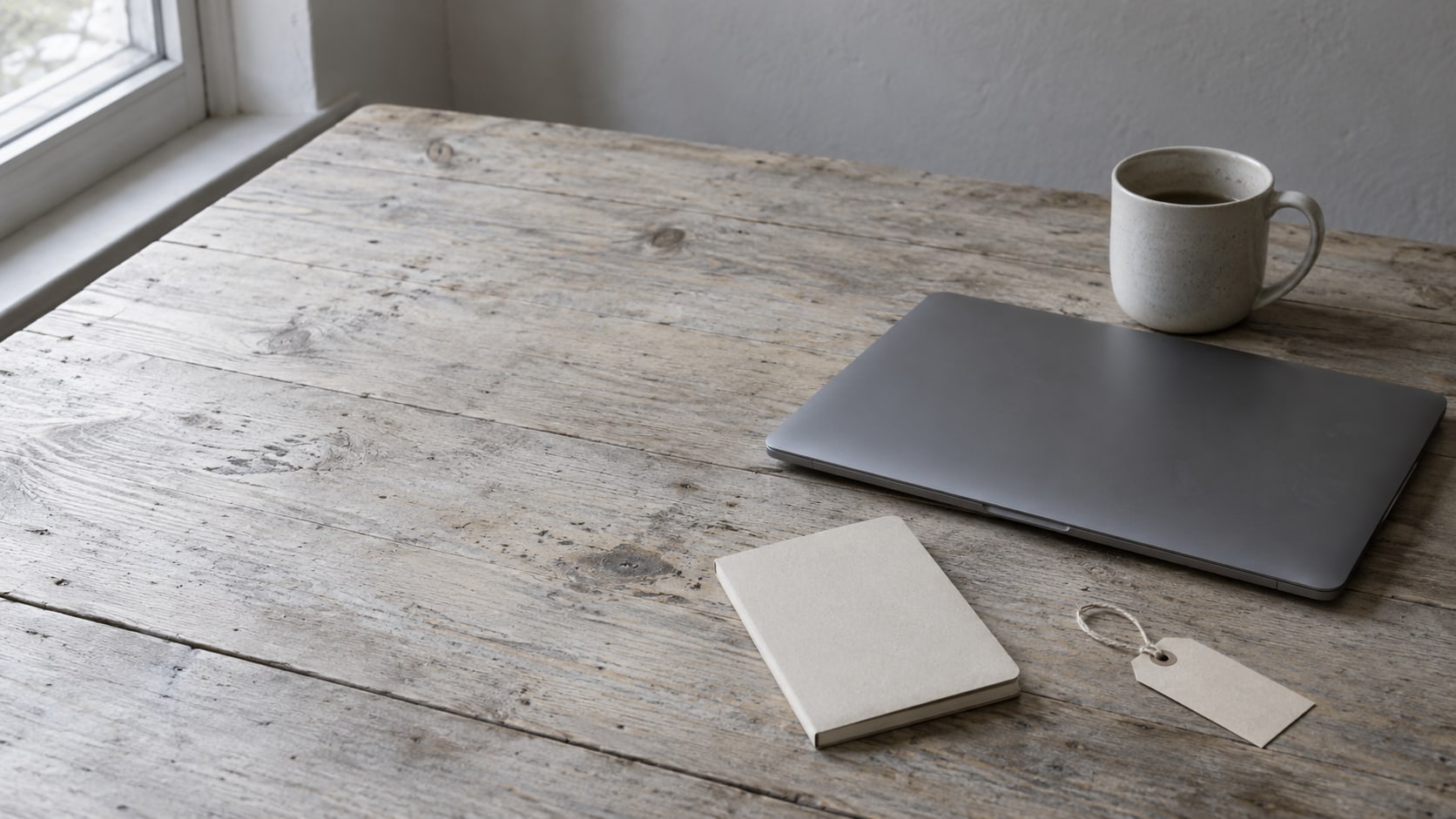 A closed laptop, a small notebook and a mug on a worn wooden desk, with a blank paper tag beside them.