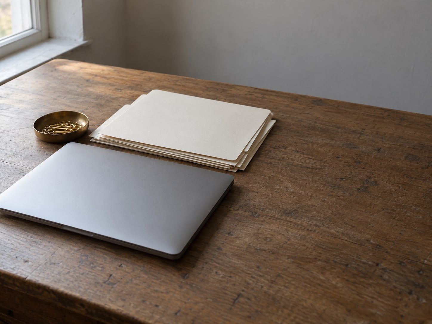 A closed laptop beside a neat stack of unmarked paper folders and a small brass paperclip dish on a plain wooden desk.