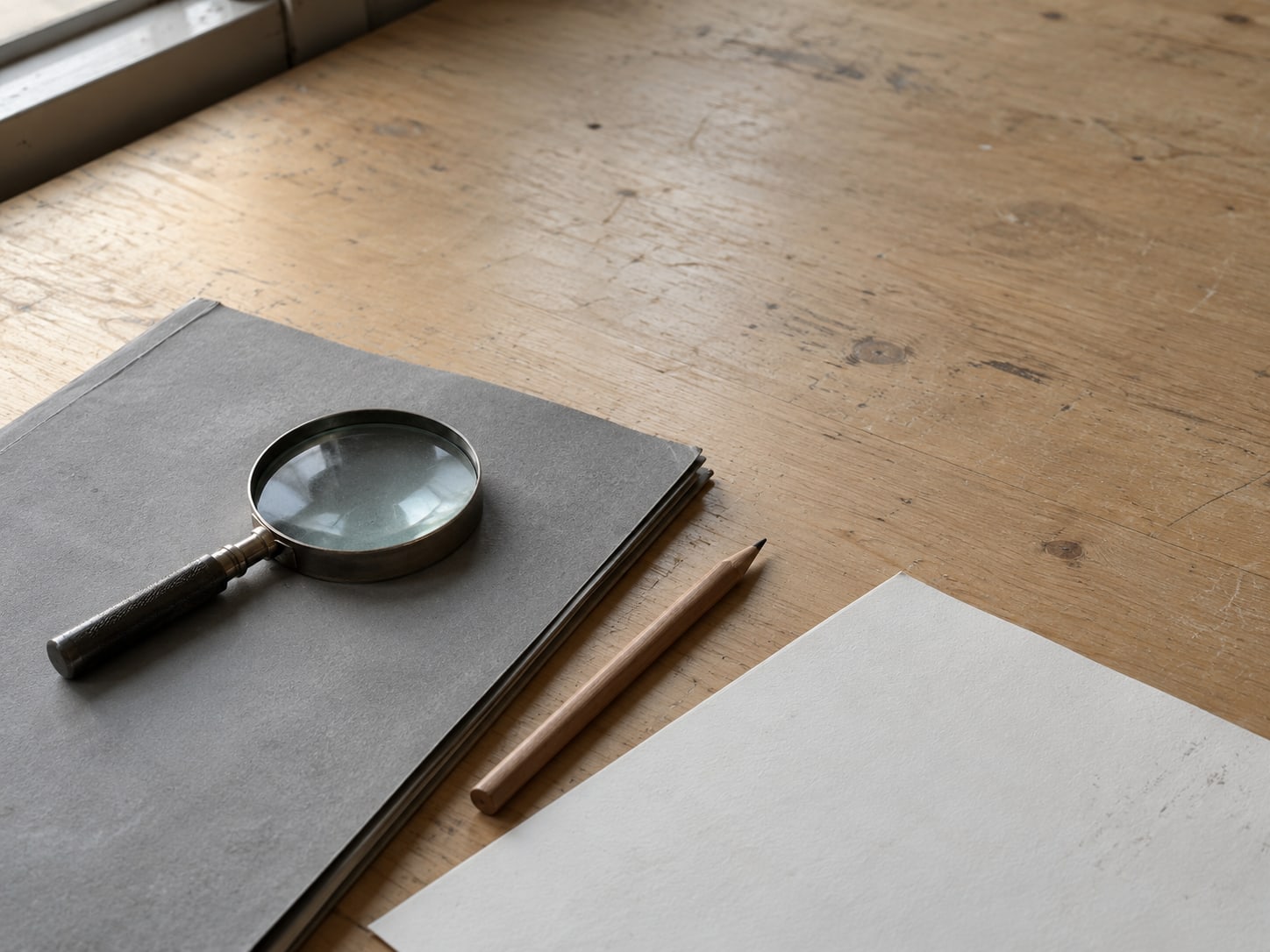 A small magnifying glass resting on a closed document folder beside a pencil with unreadable light marks on a blank sheet.