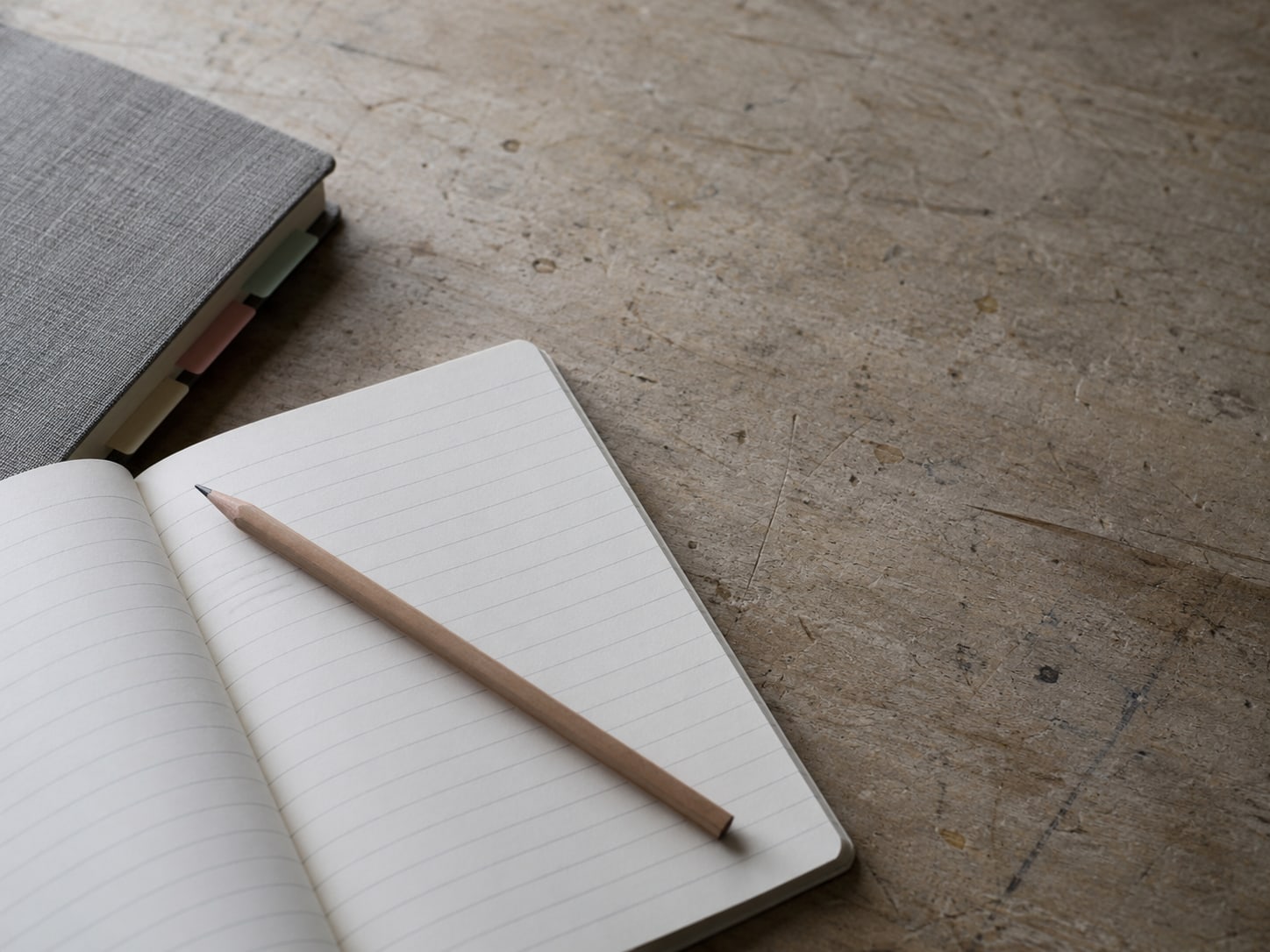 A single pencil lying across a ruled blank notepad next to a closed binder with colored index tabs on a worktable.