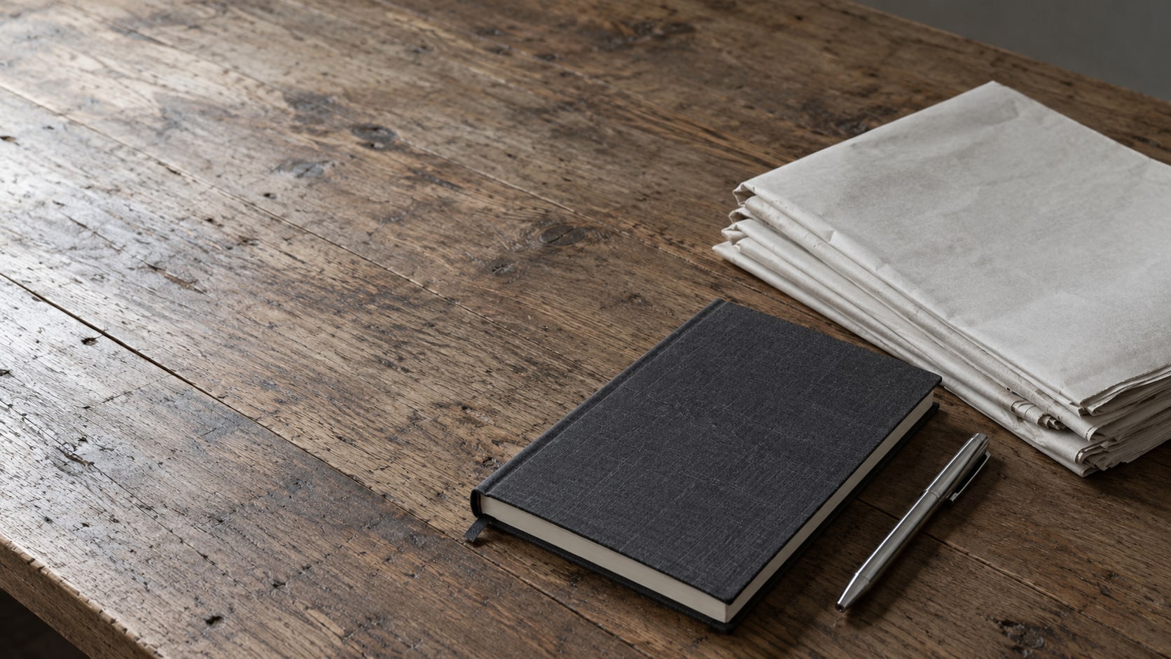Un cuaderno cerrado, un bolígrafo y una pequeña pila de páginas de periódico dobladas sobre un escritorio de madera gastada.