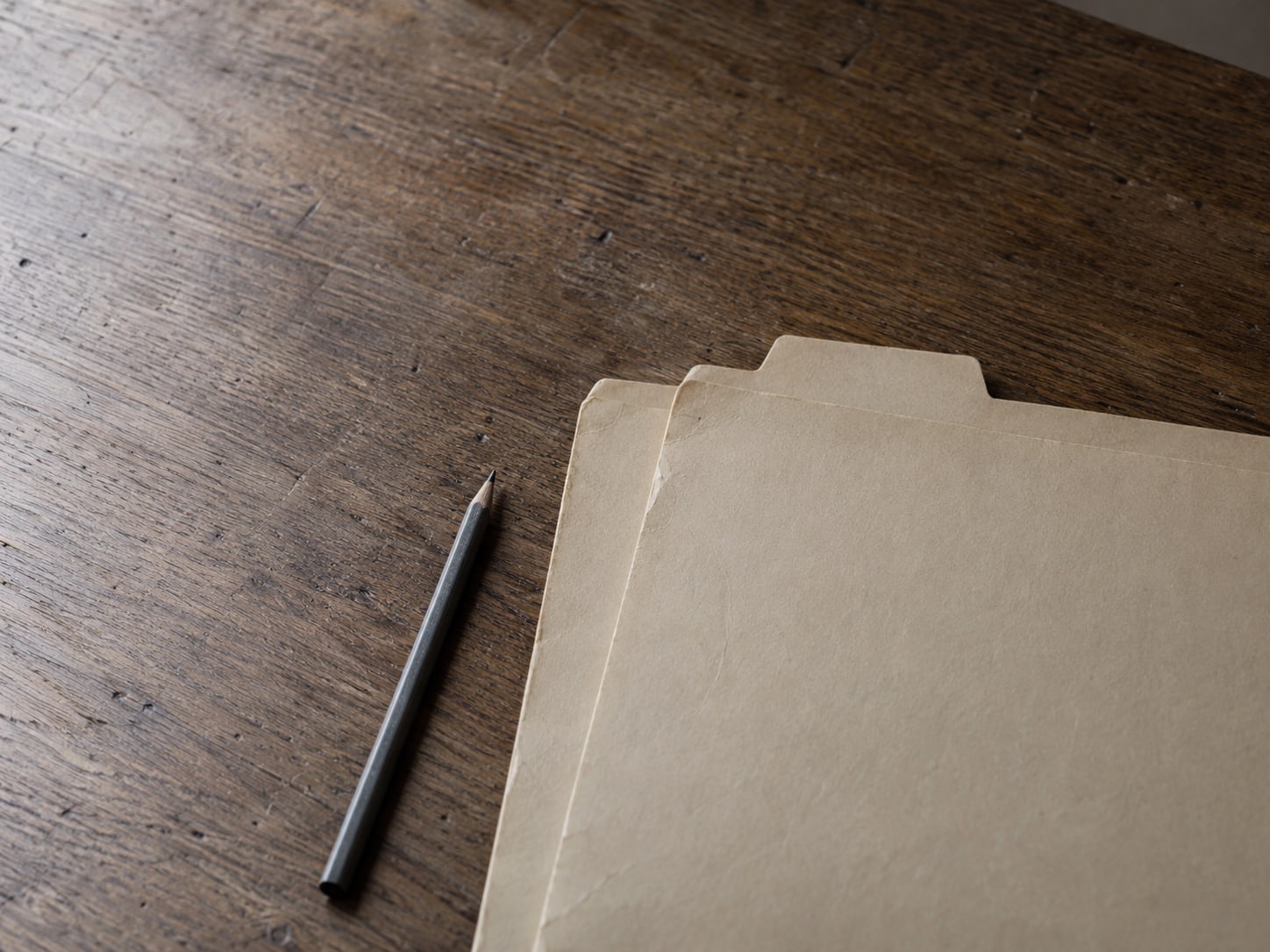 Two nearly identical manila folders sharing one tab label on a plain wooden desk, a pencil laid beside them.