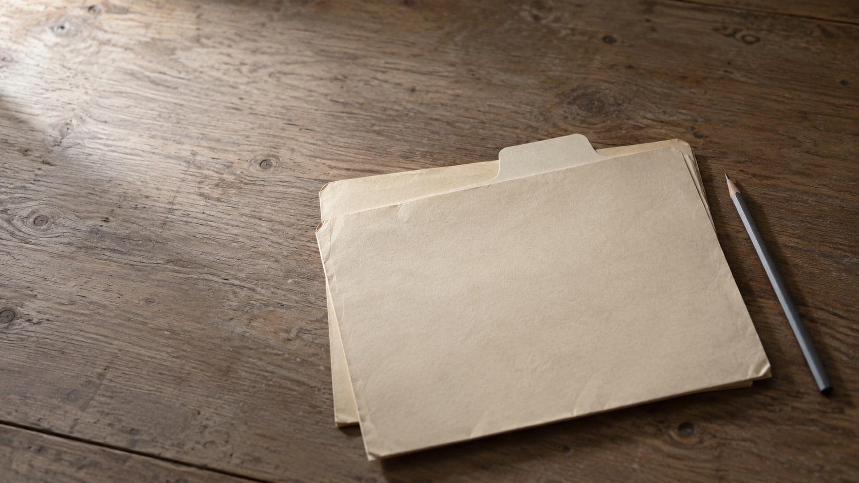 Two matching manila folders sharing one tab label on a plain wooden desk, with a pencil laid beside them.