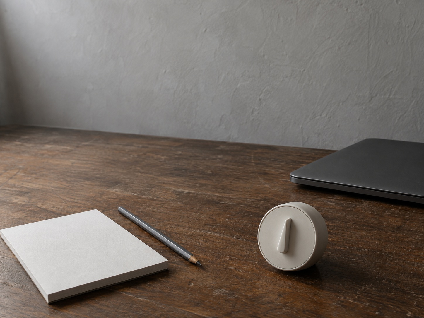 Un temporizador de cocina, un lápiz y una libreta en blanco sobre un escritorio de nogal desgastado junto a una laptop cerrada.