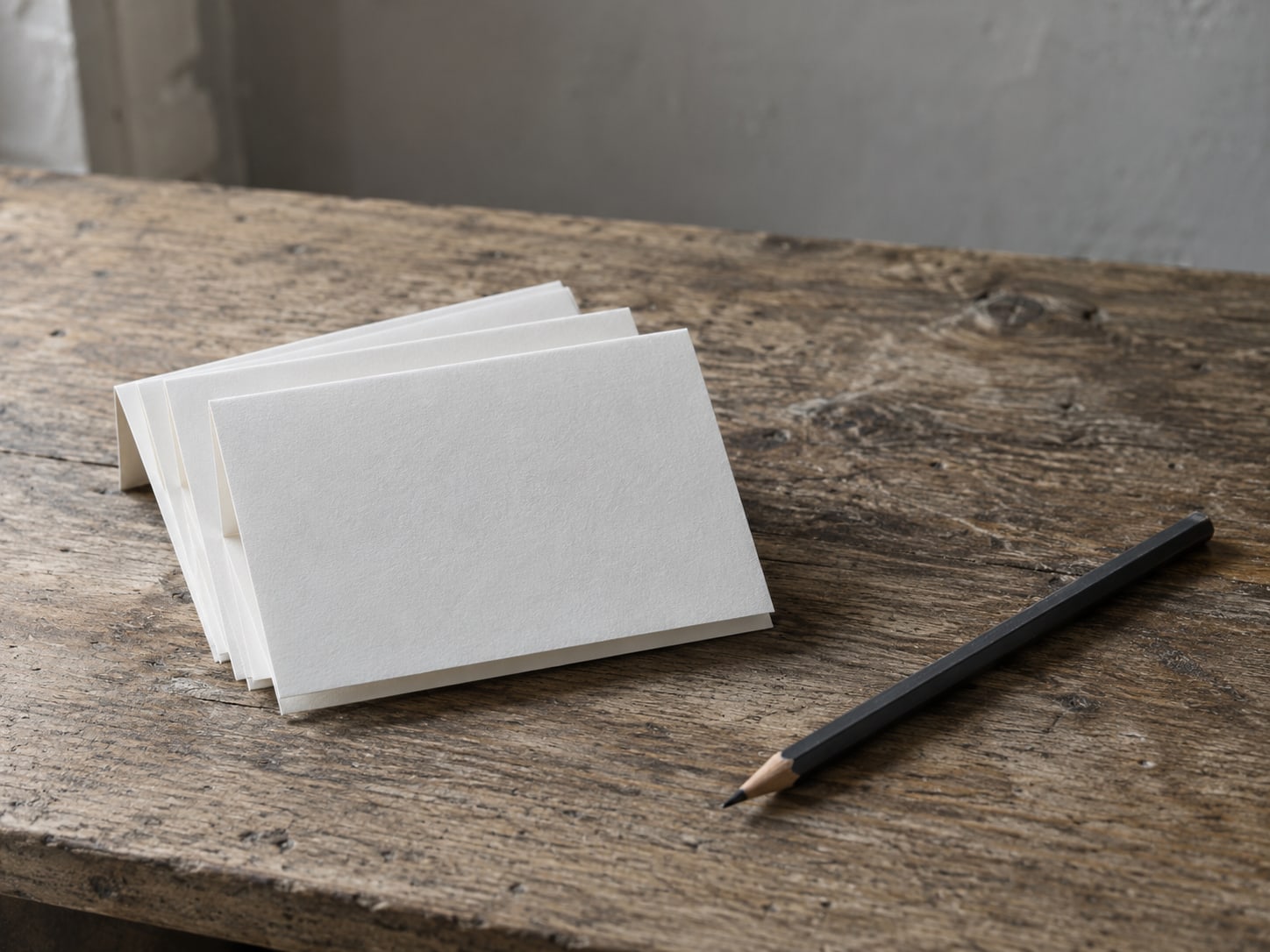A small stack of folded blank cards fanned out on a wooden table beside a pencil.