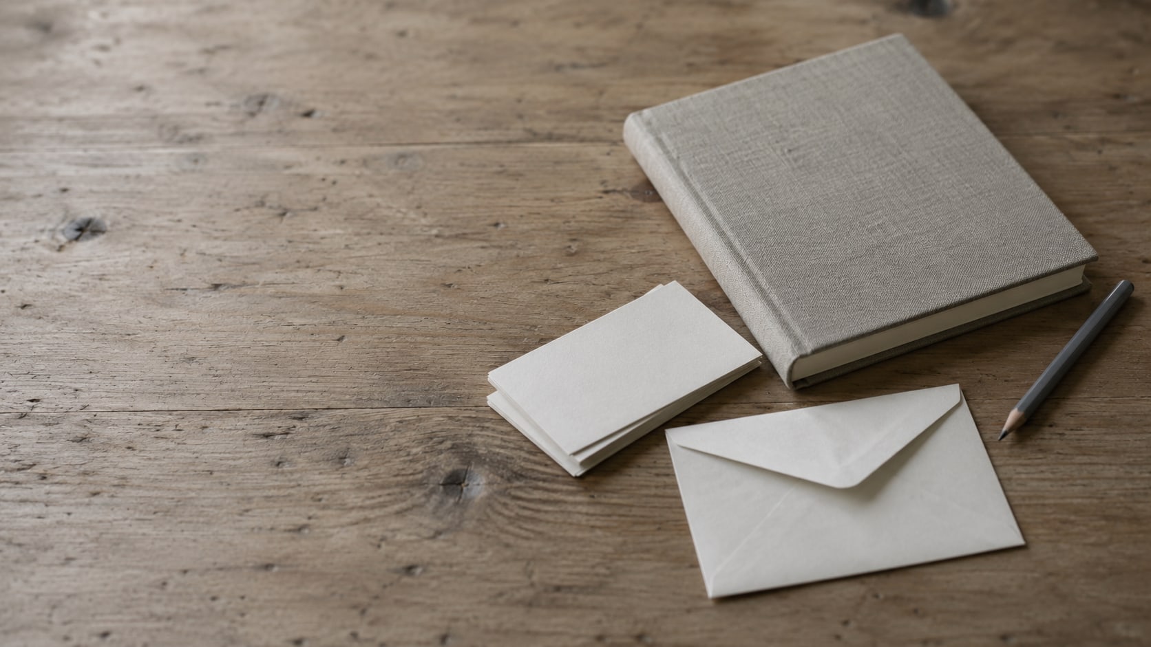 A closed notebook and a pencil on a clean wooden desk beside a small stack of blank index cards and a sealed envelope.