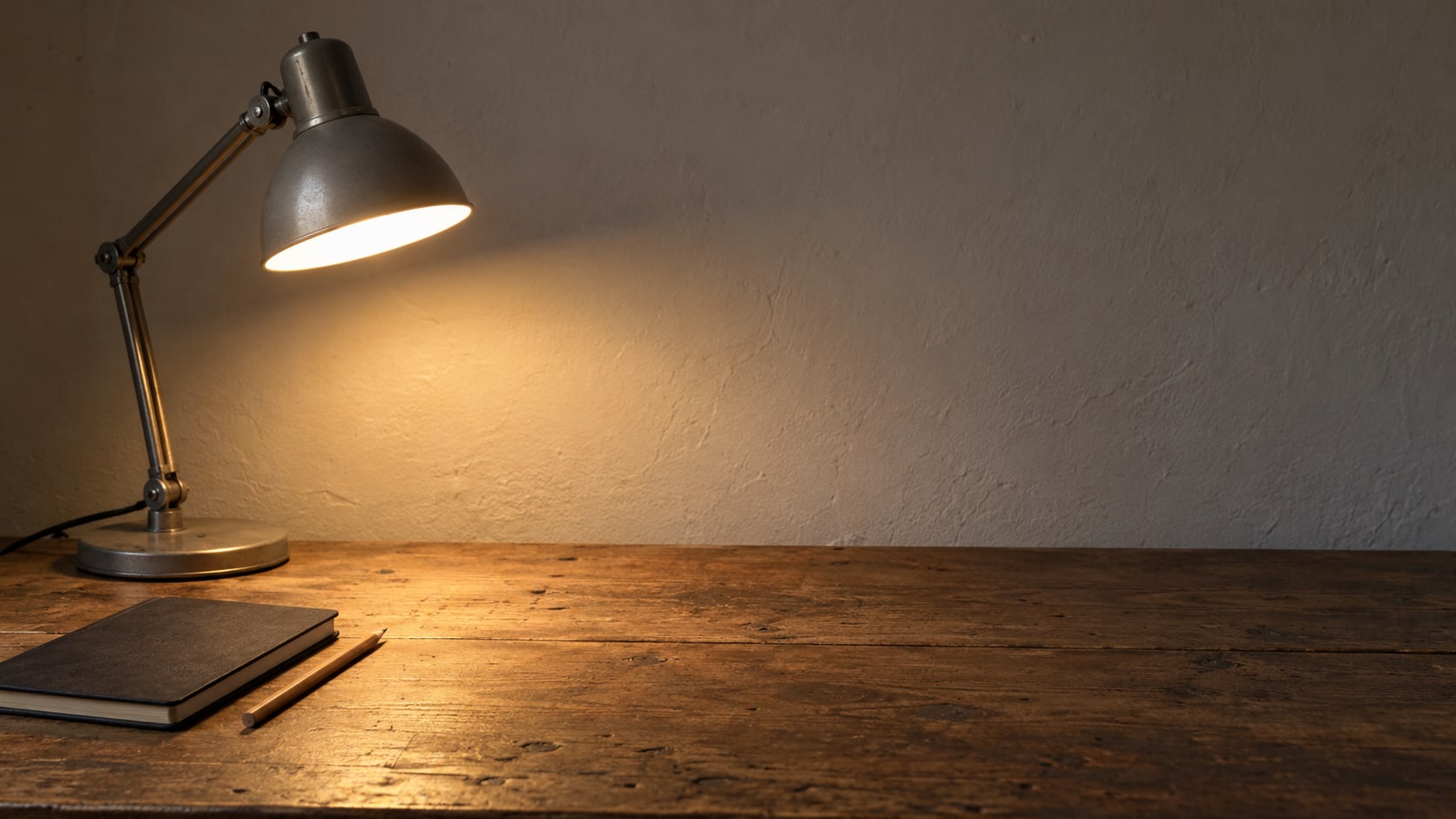 A desk lamp glowing over an empty wooden desk with a closed notebook and a pencil in the evening.