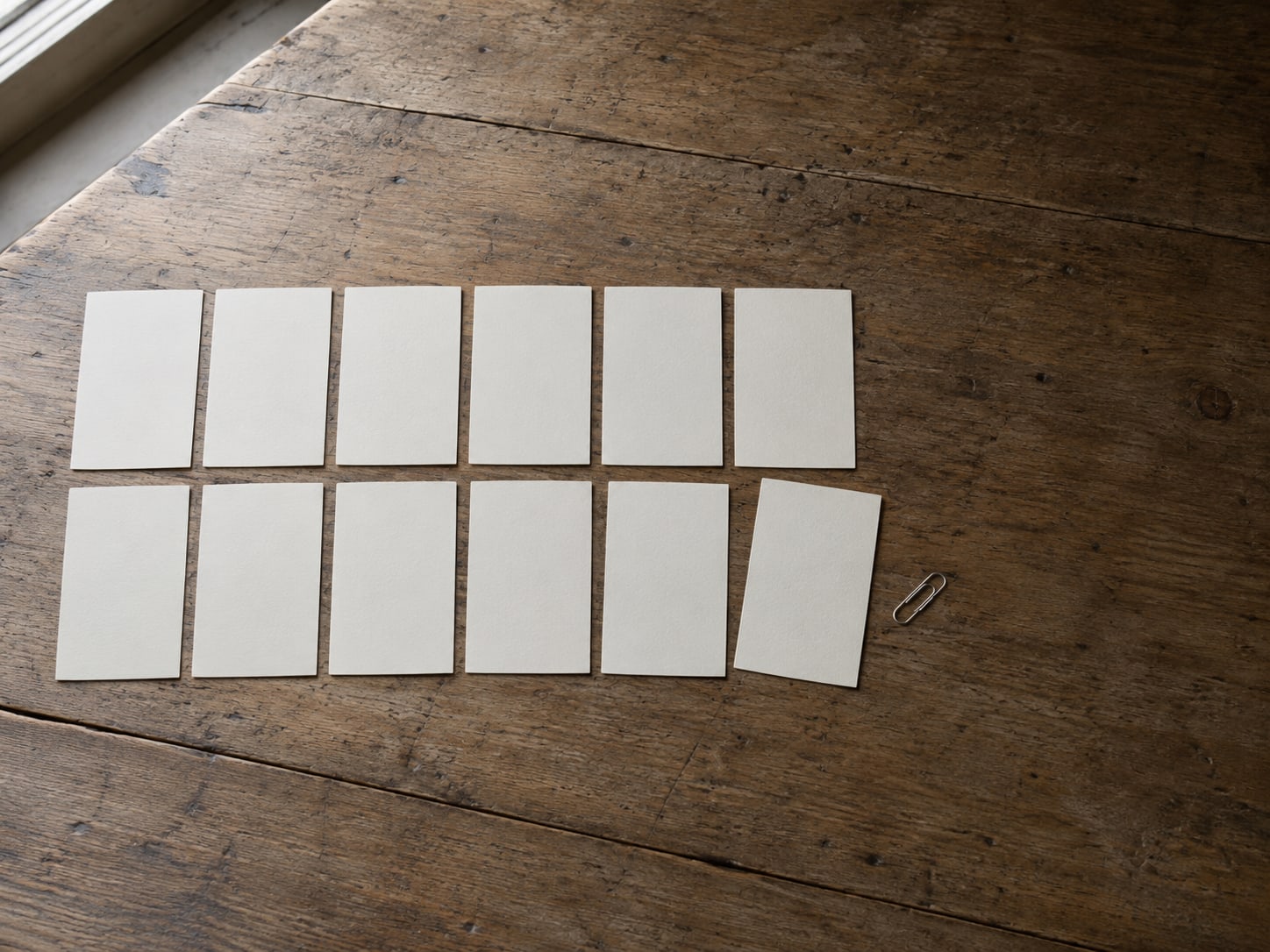 Twelve blank index cards laid in two rows on a desk with one card turned over and a small clip beside it.