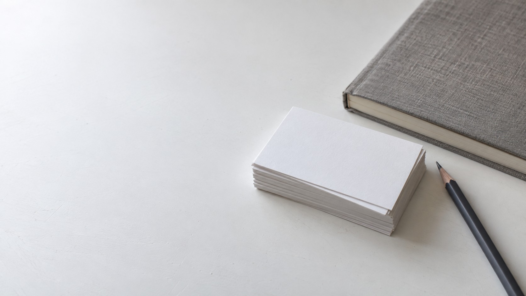 A small stack of blank business cards beside a closed sketchbook and a pencil on a white table.