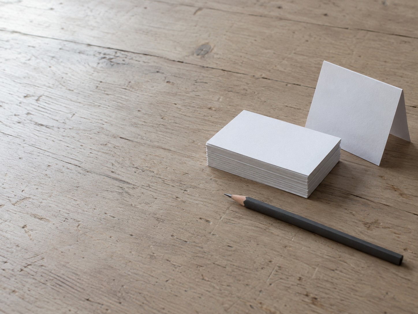 A stack of blank business cards beside a small folded paper sign and a pencil on a light wooden table.