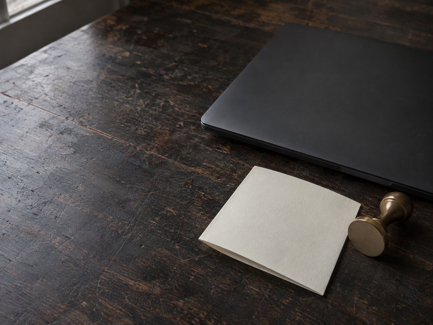A closed laptop beside a small brass stamp and a folded sheet of blank paper on a dark desk.
