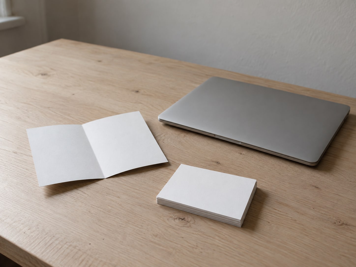 A folded paper flyer and a small stack of blank postcards resting beside a closed laptop on a light wooden desk.