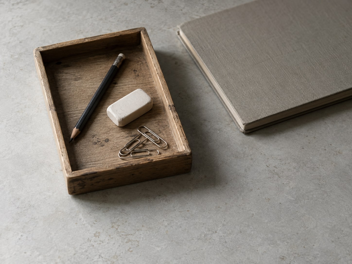 A small wooden tray holding a pencil, an eraser and a few paper clips next to a closed notebook on a desk.