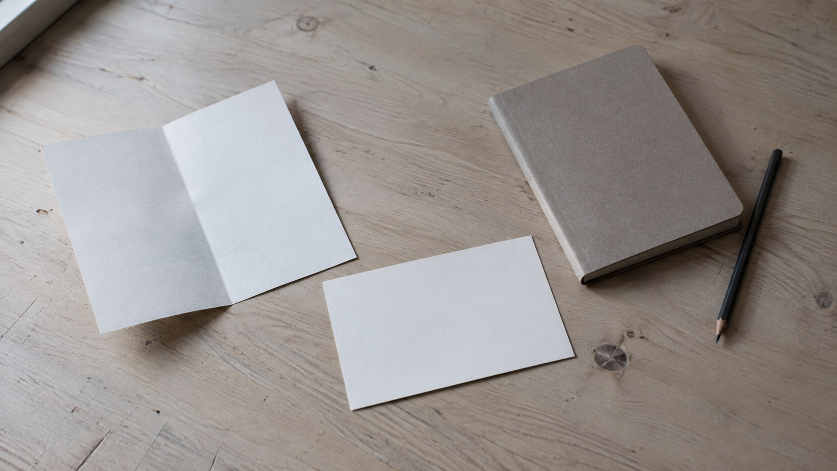 A folded paper flyer, a blank postcard and a closed notebook with a pencil arranged on a light wooden desk.