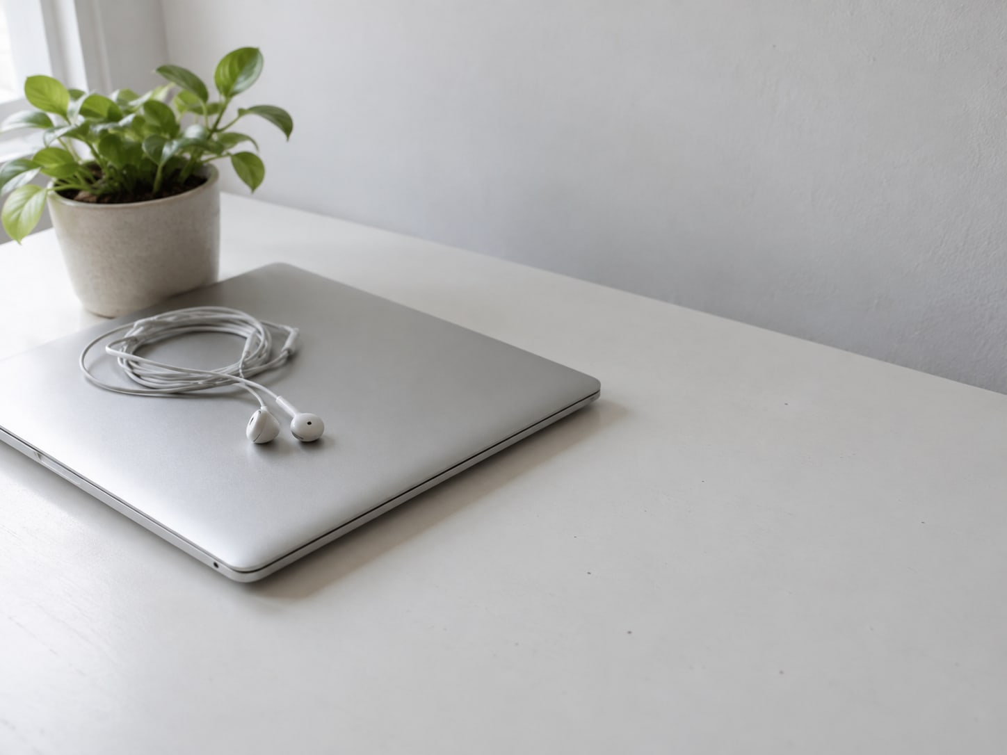 A closed laptop with a pair of wired earbuds coiled on top, next to a small potted plant on a white desk.