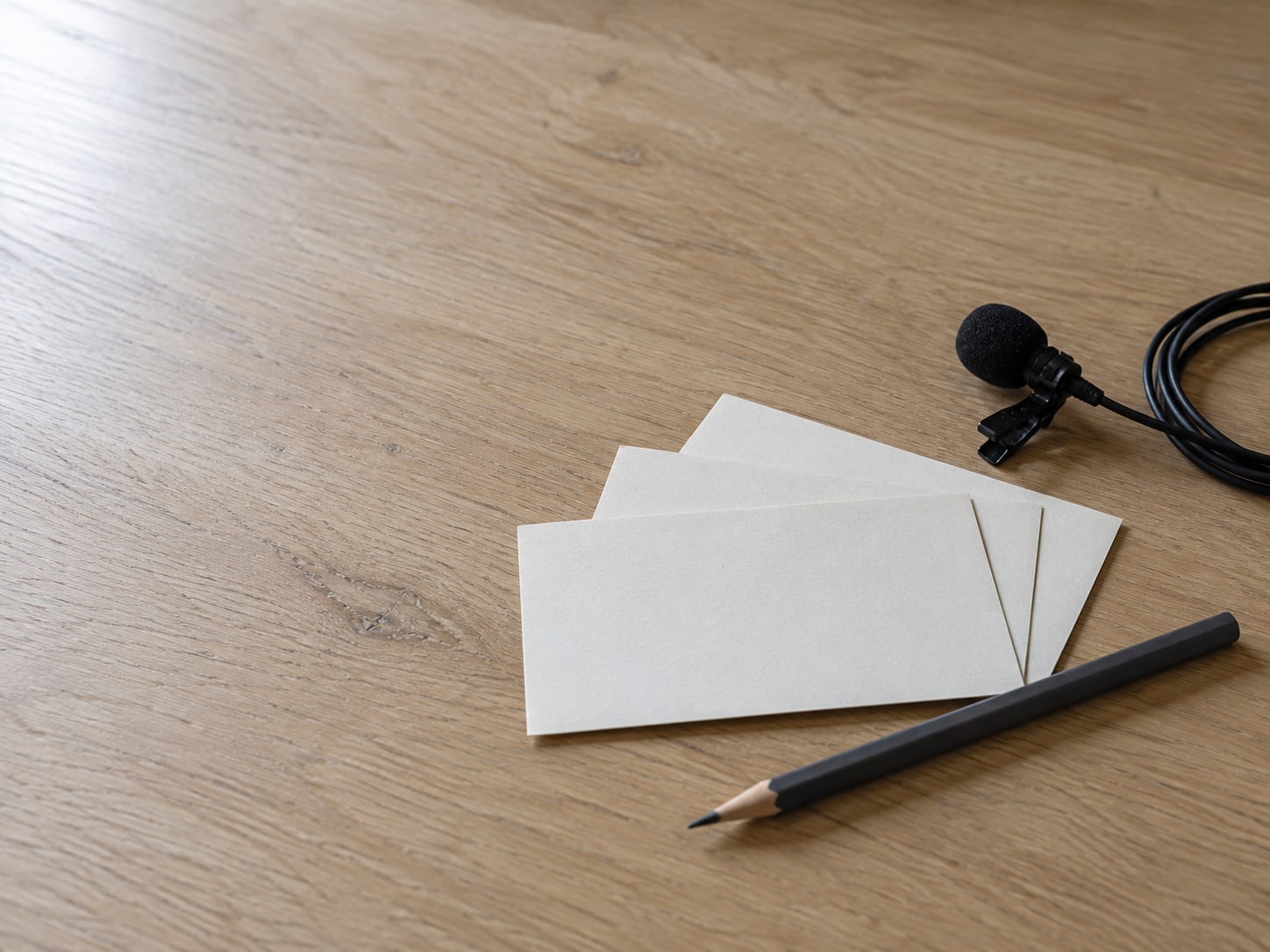 Three blank index cards fanned out beside a pencil and a small clip-on microphone on a light oak desk.
