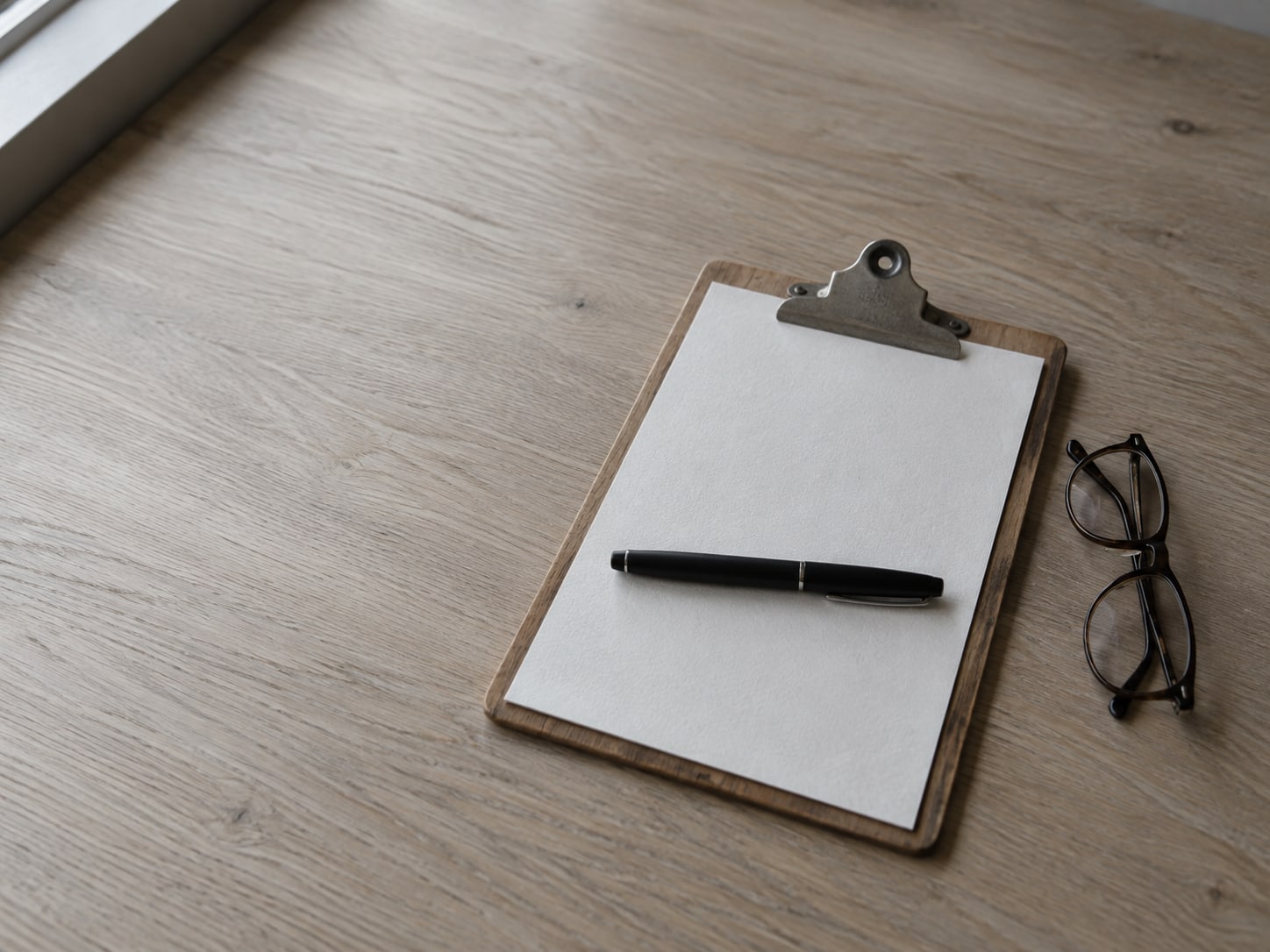 A blank sheet of paper on a clipboard with a closed pen resting across it, beside a folded pair of reading glasses on a desk.