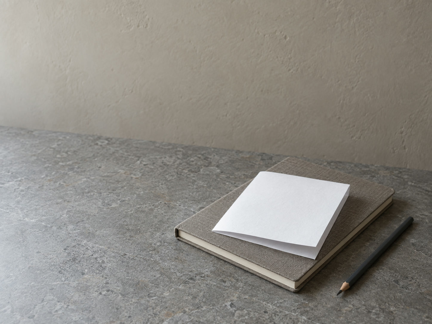 A single folded sheet of blank paper resting on a closed notebook beside a pencil on a gray stone countertop.