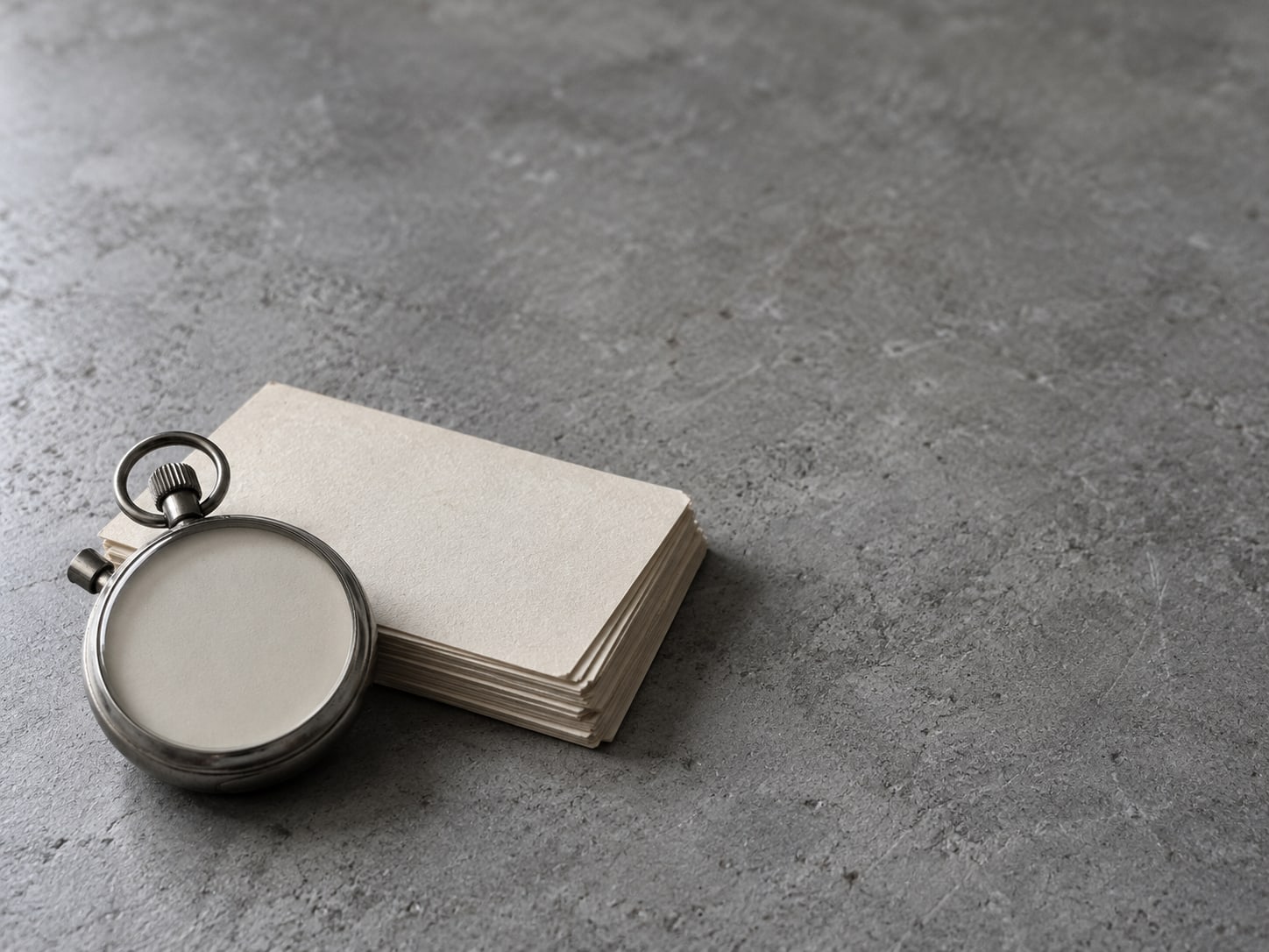 Un cronómetro acostado junto a una pequeña pila de tarjetas de índice en blanco sobre una mesa gris.