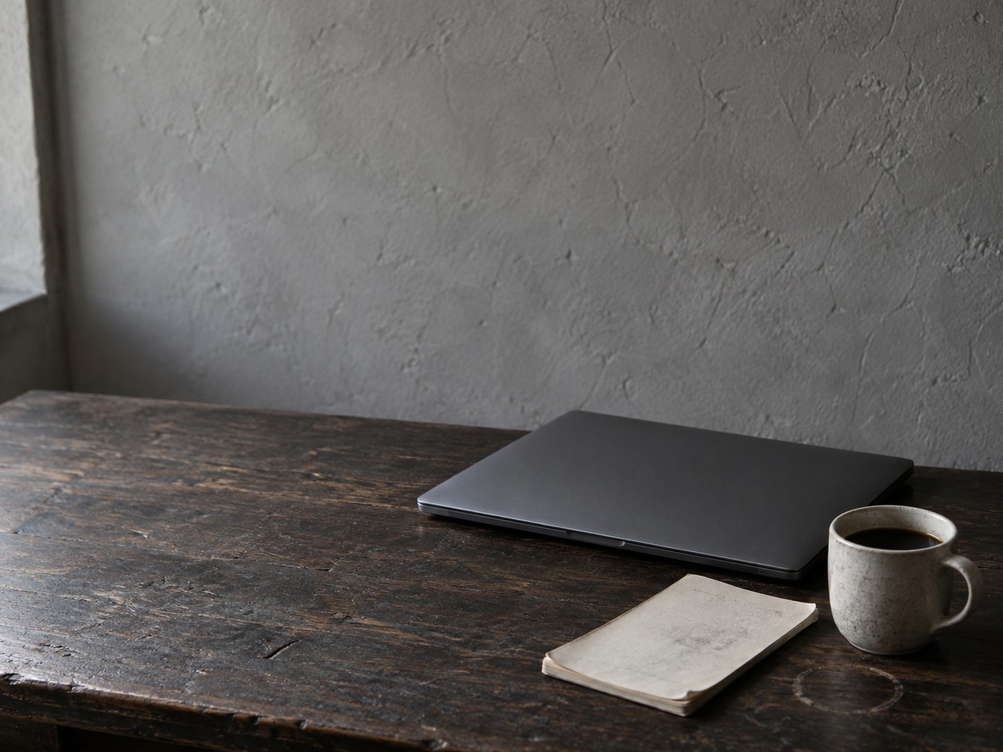 A closed laptop beside a cold cup of coffee and a notepad covered in faint pencil marks on a dark wooden desk.