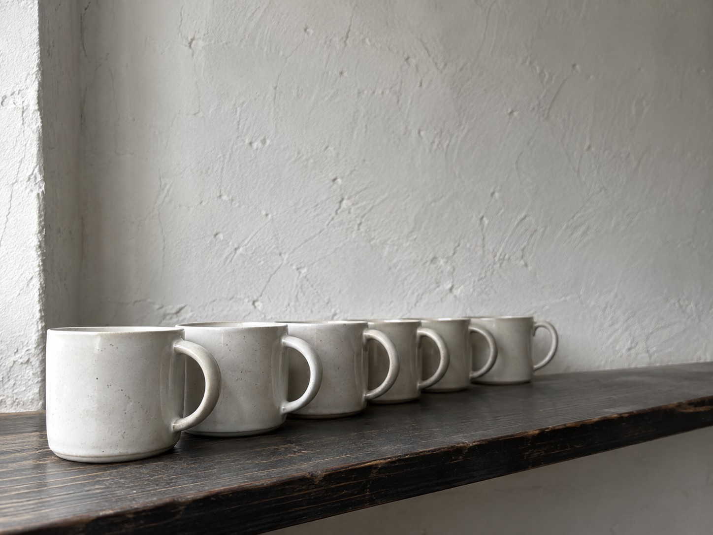 A row of identical plain white ceramic mugs lined up on a long wooden shelf against a pale wall.