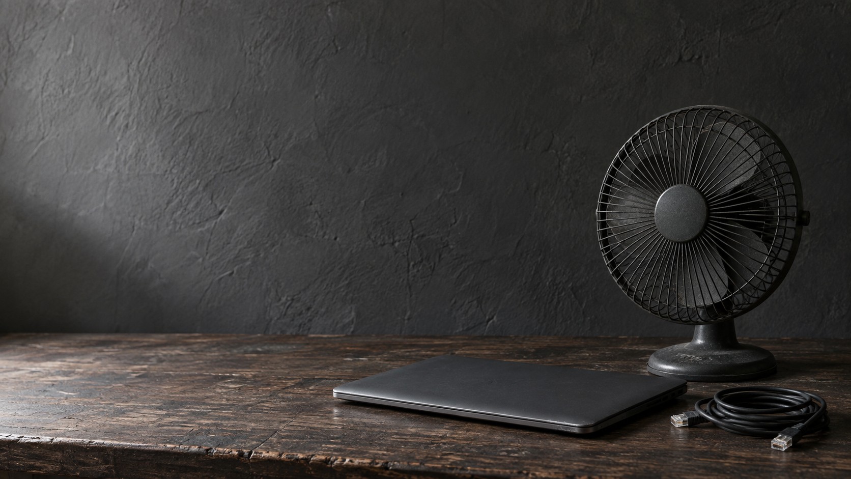 A switched-off desk fan beside a closed laptop and a coiled network cable on a dark desk.