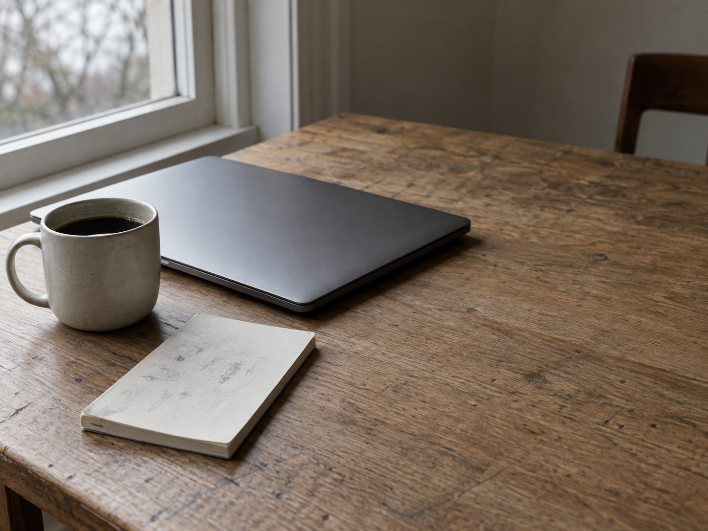 A blank notepad with faint unreadable pencil marks next to a closed laptop and a cup of coffee on a kitchen table.
