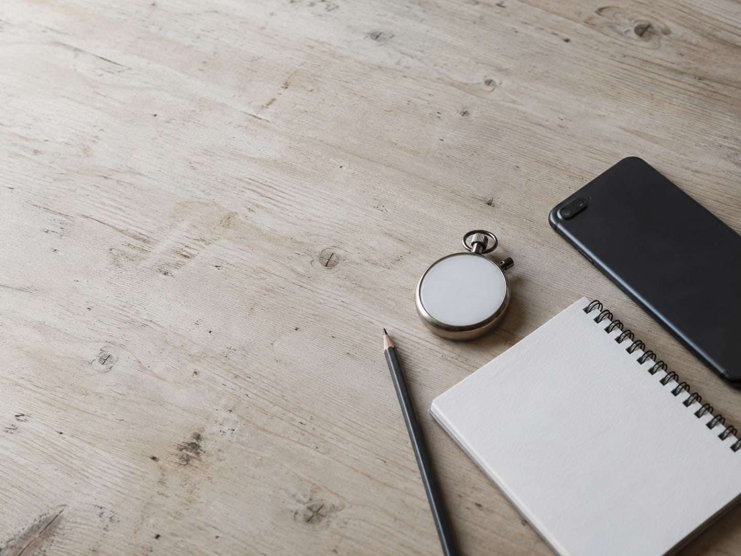 A closed smartphone lying beside a stopwatch and a blank notepad with a pencil on a pale wooden desk.
