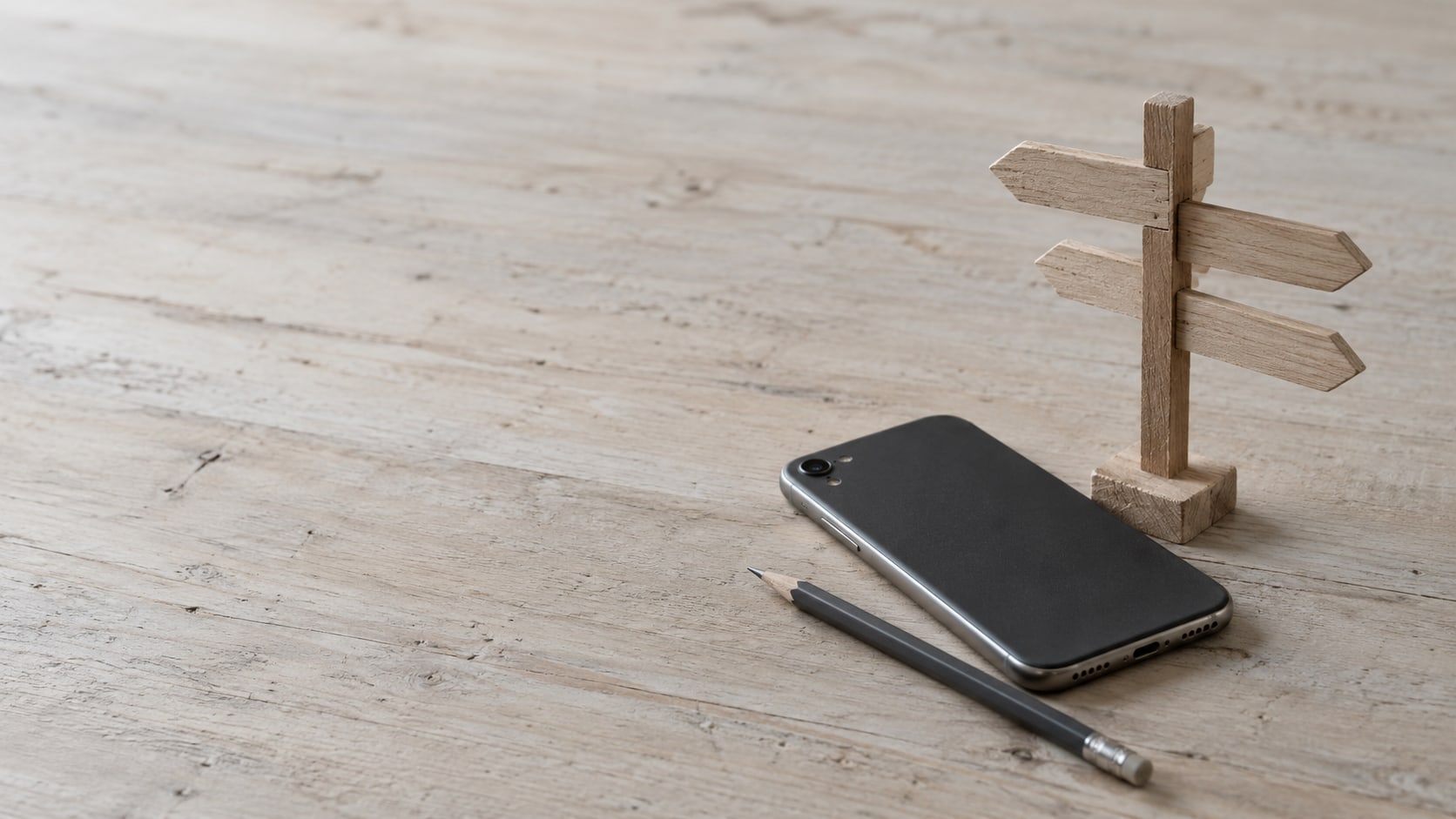 A closed smartphone and a small wooden signpost with blank arms on a pale desk beside a pencil.