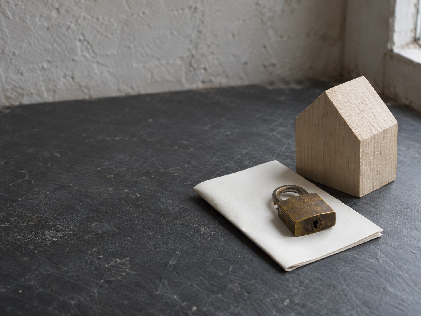 A closed brass padlock resting on a folded sheet of blank paper beside a small wooden house model on a slate desk.