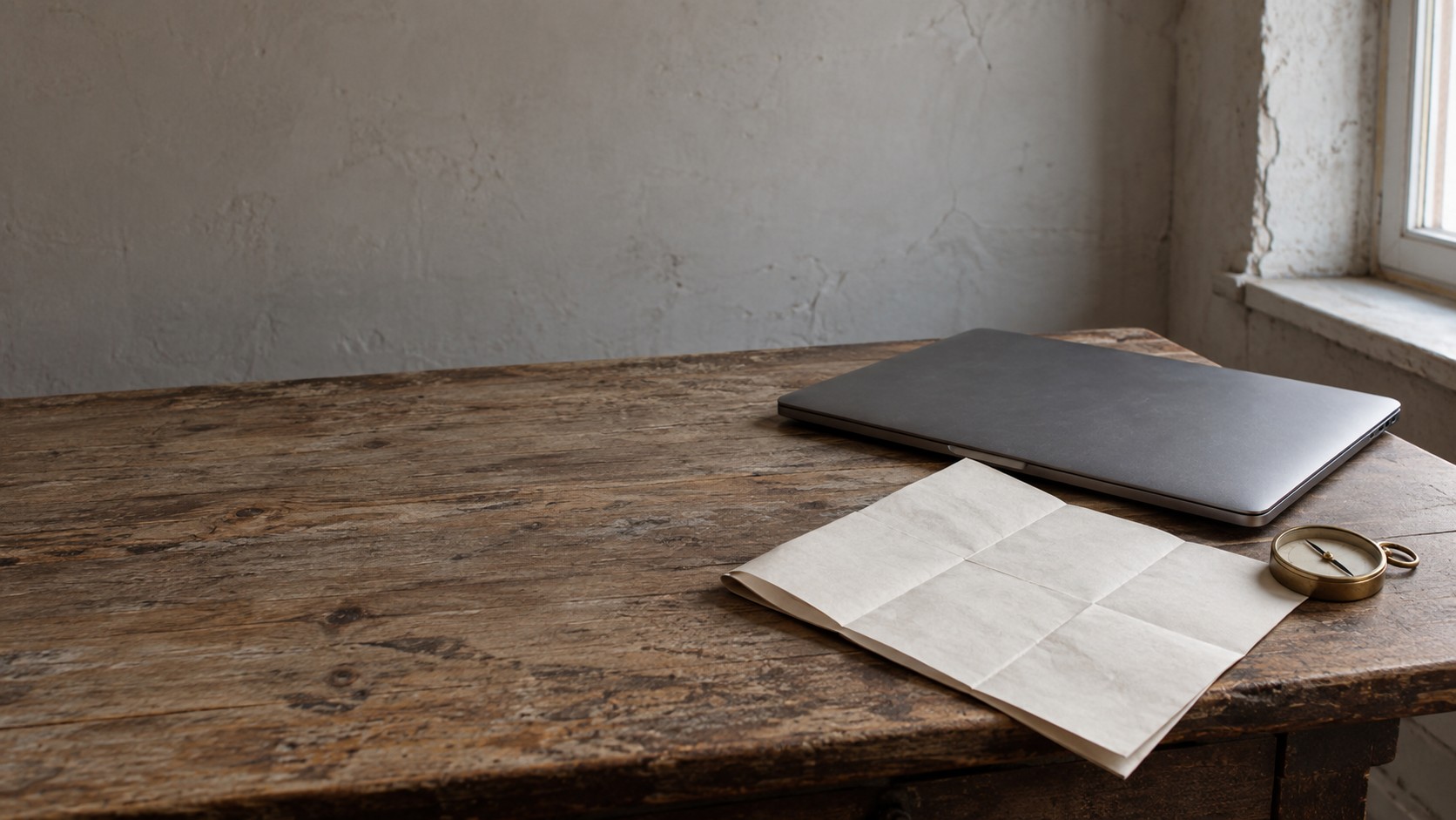 A closed laptop resting on a wooden desk beside a folded paper map and a small compass, in soft window light.