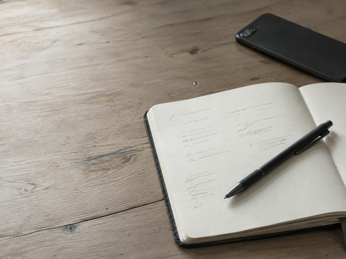 A notebook open on a desk beside a face-down phone, with short rows of unreadable pencil notes and a pen resting across the page.