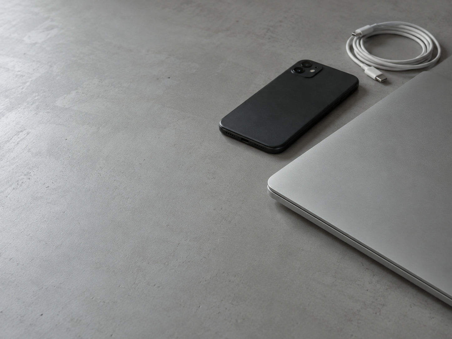 A laptop with its lid closed beside a face-down phone and a coiled charging cable on a clean gray desk.