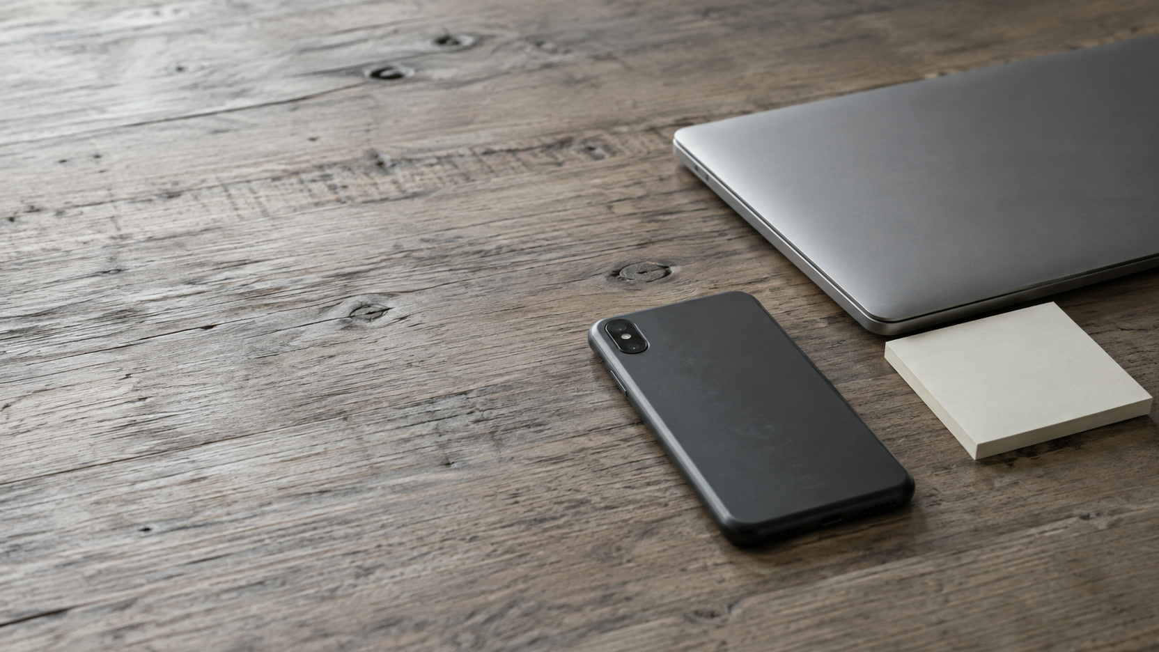 A smartphone lying face down beside a closed laptop and a blank sticky note on a wooden desk.