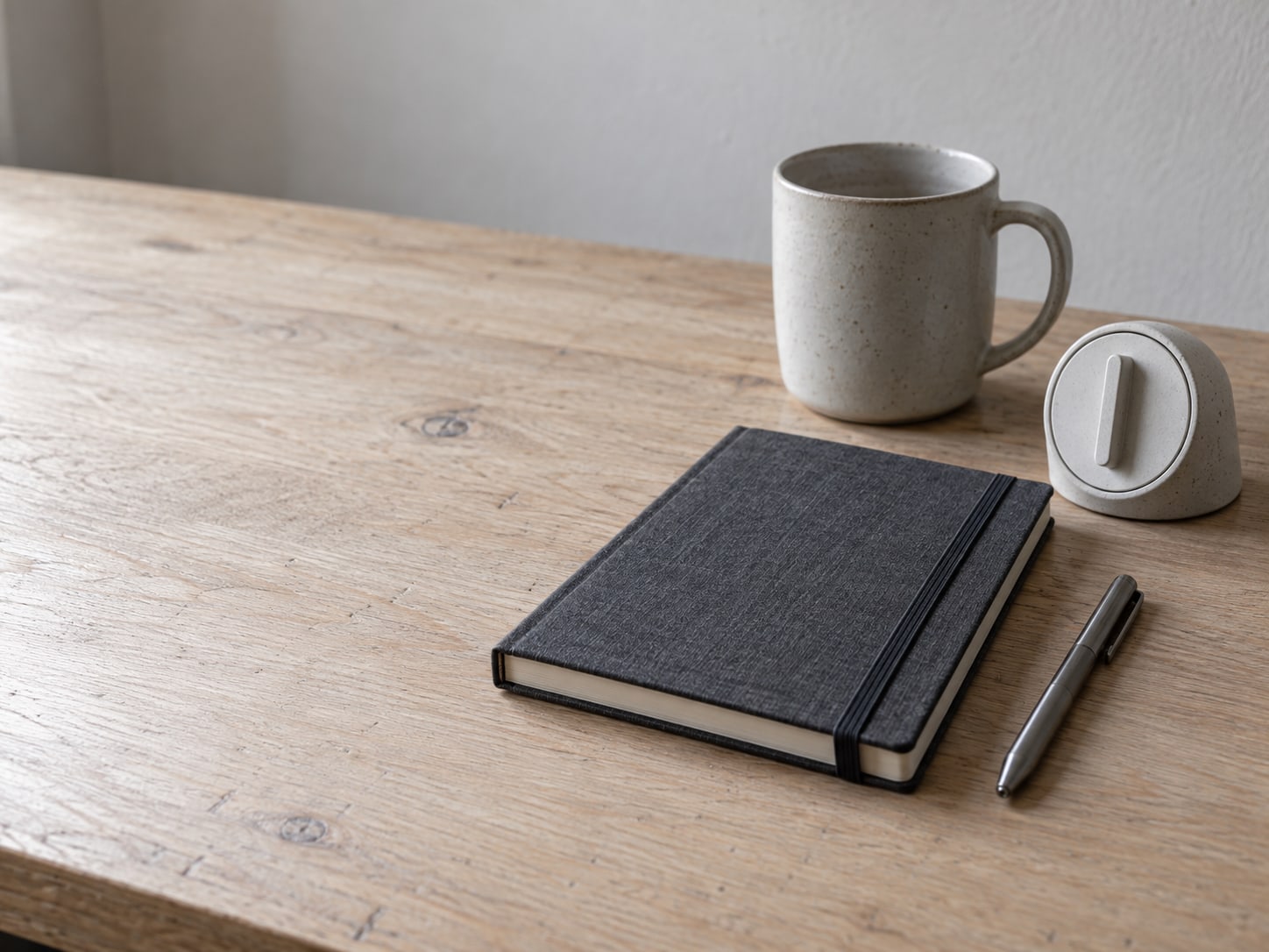 A closed notebook, a pen and a small analog kitchen timer set on a tidy desk beside a mug.