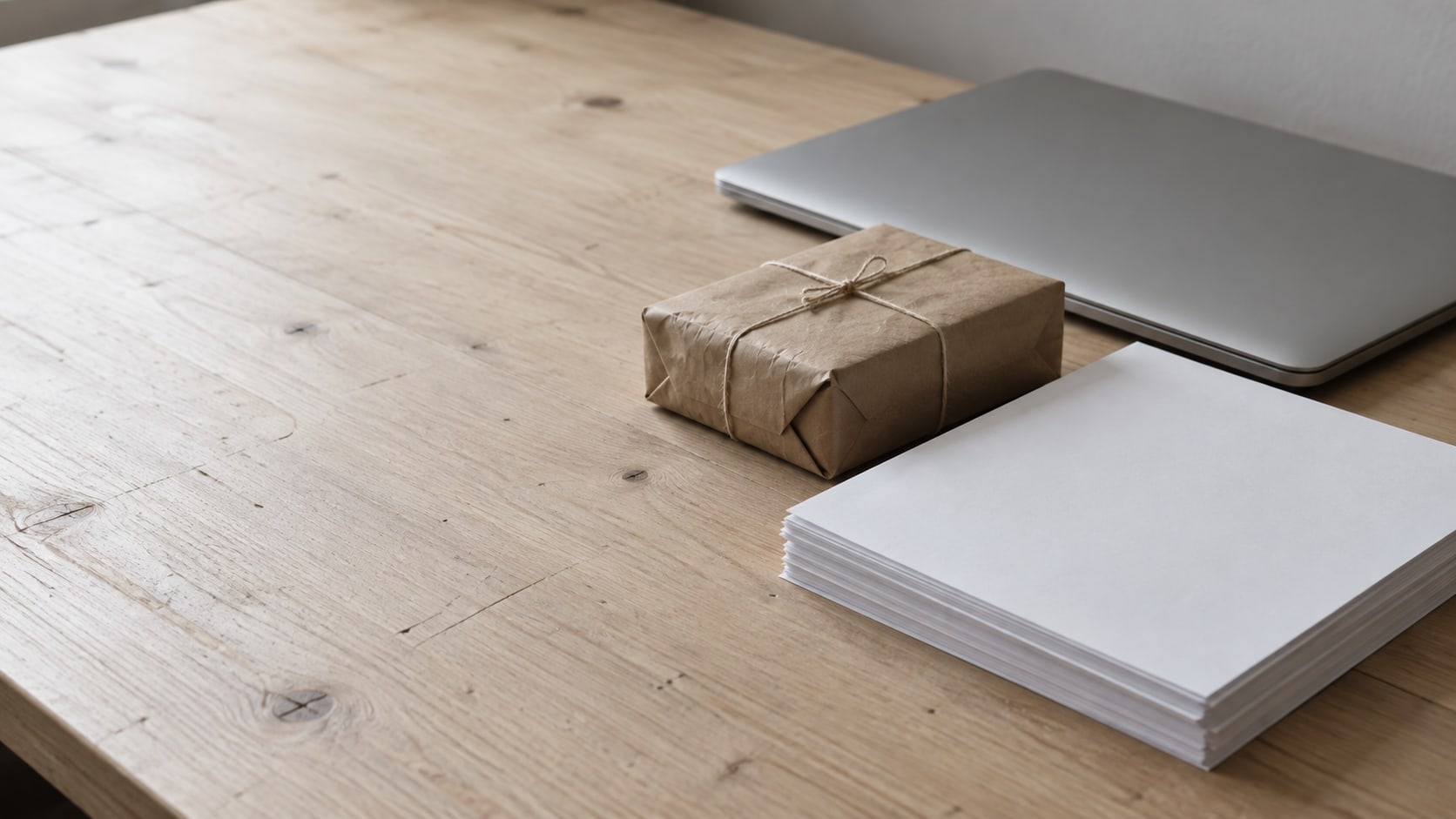 A neatly stacked set of blank paper sheets beside a small wrapped parcel and a closed laptop on a pale wooden desk.