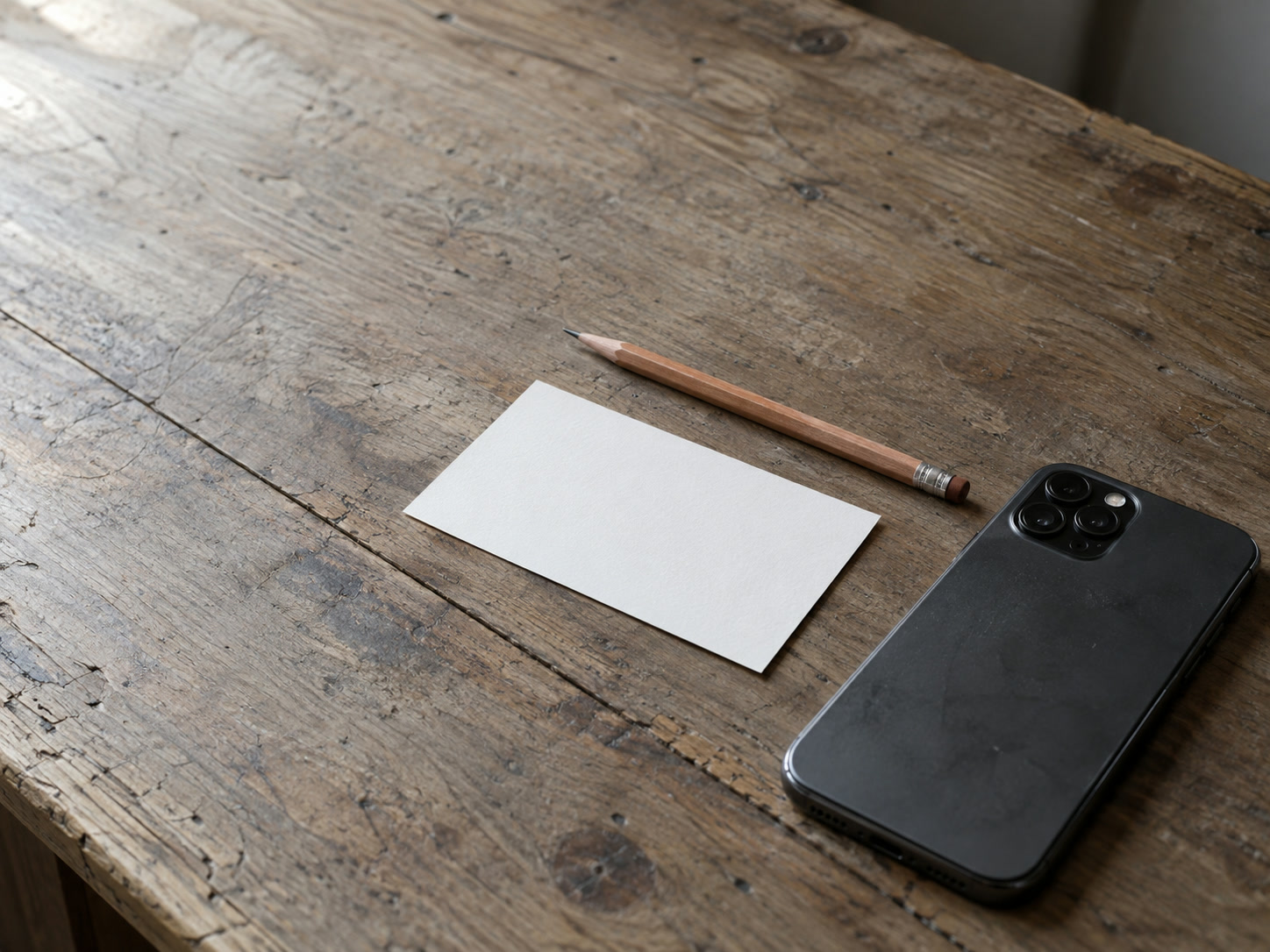 A phone lying face down beside a blank index card and a sharpened pencil on a worn wooden desk.