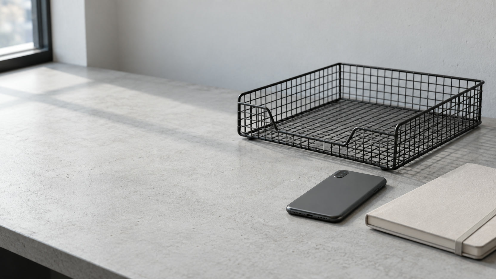 An empty wire letter tray beside a face-down phone and a closed notebook on an office desk.