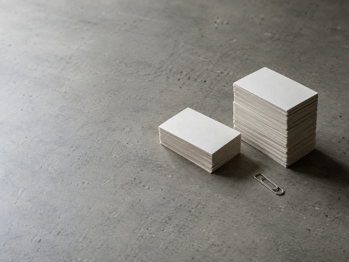 Two small stacks of blank index cards, one tall and one short, on a gray tabletop beside a single paper clip.